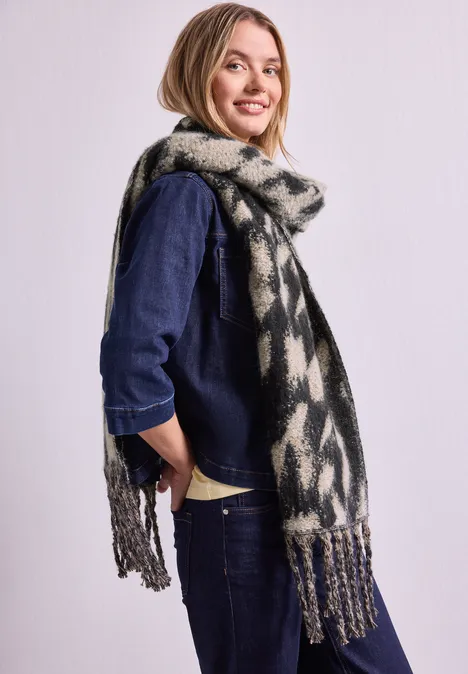 Lächelnde Frau in Denim-Outfit mit langem, gemustertem Schal vor hellem Hintergrund.