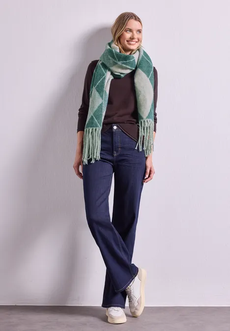 Frau in lässigem Outfit: dunkler Pullover, weite Jeans, weiße Sneaker und grüner Schal mit Muster vor neutralem Hintergrund.