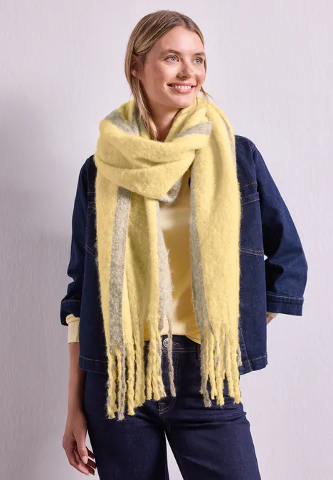Frau mit gelbem, flauschigem Schal, Jeansjacke, lächelt seitlich vor hellem Hintergrund.