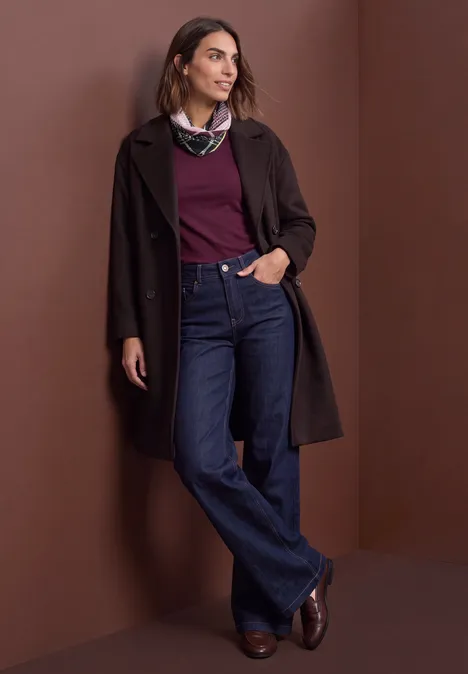 Frau lehnt an brauner Wand, trägt langen Mantel, weinroten Pullover, weites Jeans und braune Stiefeletten.