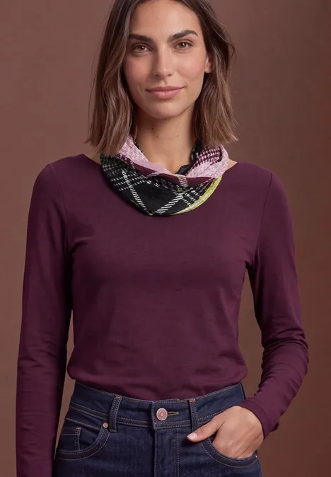 Frau im violetten Langarmshirt und kariertem Halstuch vor braunem Hintergrund, Hand locker in der Jeanshose