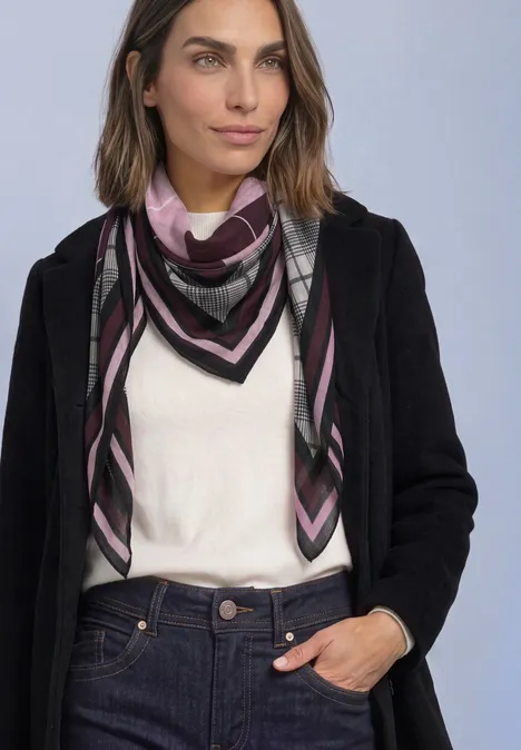 Sjaal met argyle-patroon berry plum Frau mit schwarzem Mantel, weißem Oberteil, gemustertem Schal und Jeans vor neutralem Hintergrund.