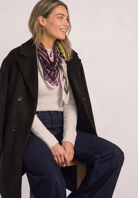 Doek met grafisch patroon multicolor Eine lächelnde Frau sitzt seitlich, trägt dunklen Mantel, weißen Pullover, Halstuch und weite Jeans.