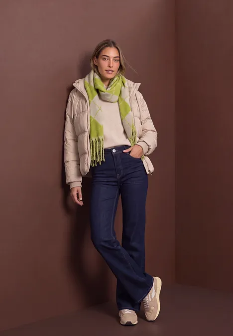 Eine Frau lehnt in beiger Steppjacke, grüner Strickschal und weiten Jeans vor braunem Hintergrund.