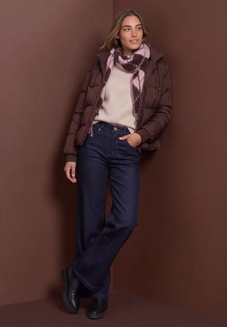 Frau in brauner Steppjacke, weißem Pullover, Jeans und Schal vor braunem Hintergrund.