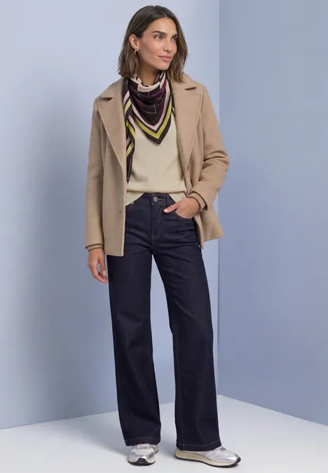 Frau in beiger Jacke, cremeweißem Pullover, farbigem Tuch, weiter Jeans und weißen Sneakern.