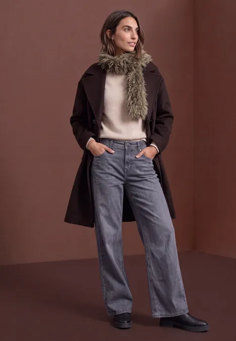 Schal aus Fake Fur in Unifarbe oatly sand Frau vor braunem Hintergrund, trägt dunkelbraunen Mantel, hellen Pullover, weite graue Hose, grünen Schal und Stiefel.