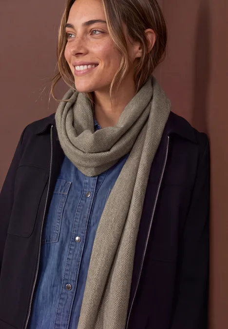 Geplooide sjaal met visgraatpatroon bark brown Frau mit schwarzem Mantel, blauem Jeanshemd und beigem Schal lächelt zur Seite.