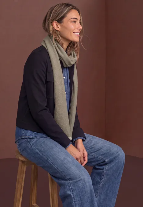 Geplooide sjaal met visgraatpatroon bark brown Frau sitzt auf Hocker, trägt Jeans, Schal und Jacke, lächelt seitlich