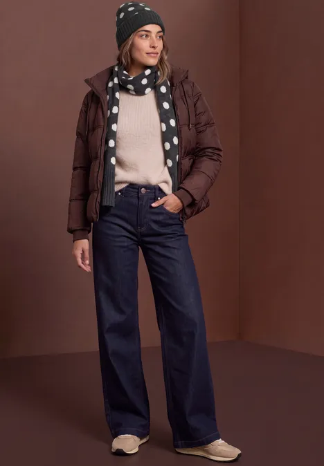 Frau in brauner Daunenjacke, gepunktetem Schal & Mütze, hellem Pullover, weiten Jeans und Sneakers.