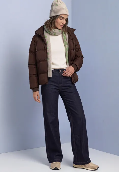 Muts met stippenpatroon clay sand mel. Vrouw met bruine pufferjas, witte trui, wijde jeans en muts, voor een lichtblauwe achtergrond.