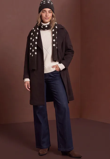 Frau in dunklem Mantel, weißem Pullover, blauen Jeans, gemusterter Mütze und Schal vor braunem Hintergrund.