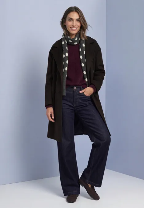 Longue écharpe à motifs de pois armor grey mel. Eine lächelnde Frau in langem Mantel, dunklen Jeans und Polka-Punkt-Schal vor neutralem Hintergrund.