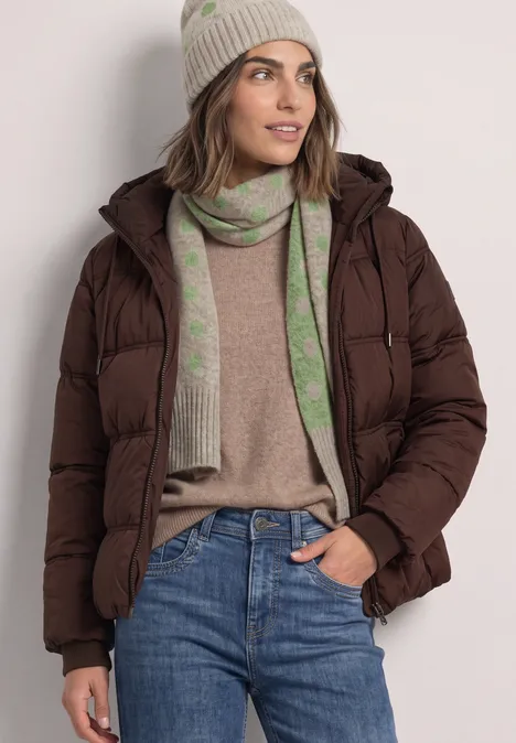 Lange sjaal met stippenpatroon clay sand mel. Vrouw in bruine donsjas, beige muts, groene sjaal, beige trui en blauwe jeans.