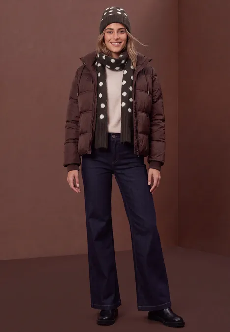 Longue écharpe à motifs de pois berry plum mel. Lächelnde Frau in brauner Daunenjacke, gepunktetem Schal, Mütze und dunklen Jeans vor braunem Hintergrund.