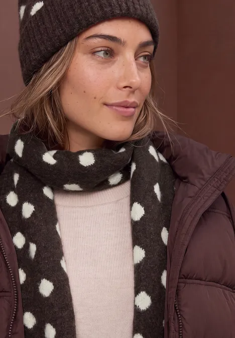 Longue écharpe à motifs de pois berry plum mel. Frau mit brauner Mütze und gepunktetem Schal, beigem Pullover und brauner Daunenjacke.