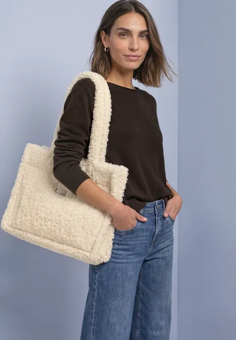 Teddy-draagtas in effen kleur barley beige Vrouw in donkerbruin trui en spijkerbroek met een zachte crèmekleurige shopper voor een lichte achtergrond.