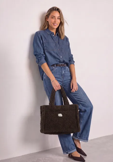 Frau in Jeanshemd und Jeanshose mit schwarzer, flauschiger Tasche vor hellem Hintergrund.