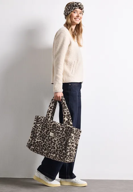 Teddy-Tragetasche mit Leo-Muster auburn brown Frau mit Leopardmuster-Mütze, beigem Pullover, weiten Jeans und gelben Schuhen trägt Leoparden-Tasche.