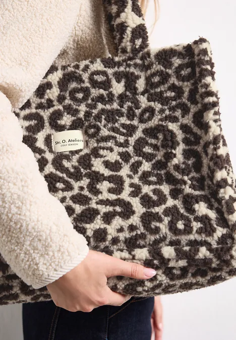 Teddy-Tragetasche mit Leo-Muster auburn brown Eine flauschige Leoparden-Tasche aus Teddyfell wird von einer Hand gehalten.