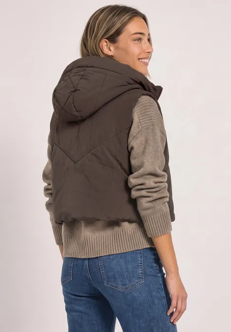 Eine Frau von hinten, die eine braune Steppweste mit Kapuze und Jeans trägt und seitlich lächelt.