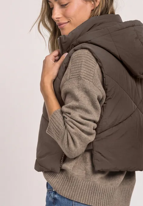 Femme en doudoune marron à capuche sur pull en maille beige, vue de profil, le regard vers le bas.