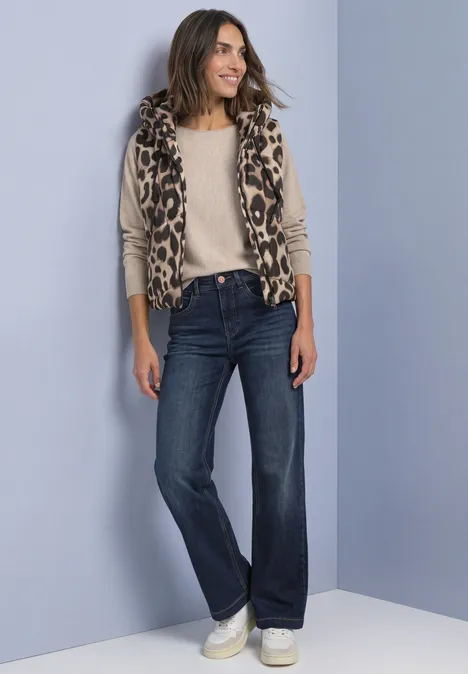 Vrouw in casual outfit: luipaardvest, licht shirt, wijde jeans en witte sneakers voor grijze achtergrond.