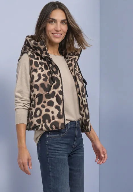 Vrouw draagt mouwloos leopardprint bodywarmer over een beige trui en jeans, glimlacht.