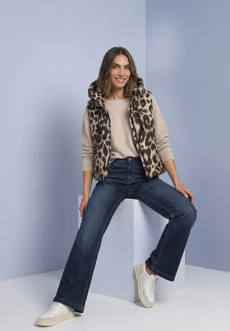 Vrouw in luipaardbodywarmer, beige top en flared jeans zit ontspannen op een witte kubus met blauwe achtergrond.