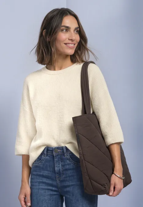 Frau mit cremefarbenem Pullover, blauen Jeans und brauner Stepp-Tasche, lächelnd vor neutralem Hintergrund.