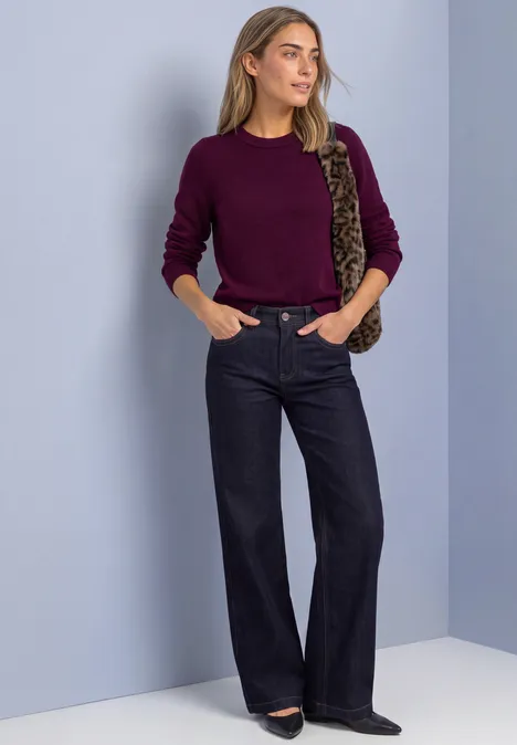 Une femme devant un mur clair porte un pull bordeaux, un jean large foncé et un sac imprimé léopard.