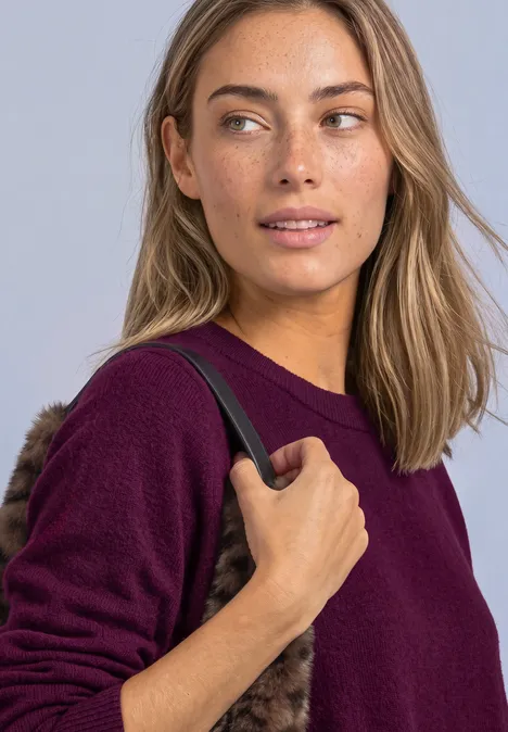 Femme aux taches de rousseur et cheveux mi-longs, porte un pull violet et tient la anse d’un sac noir.