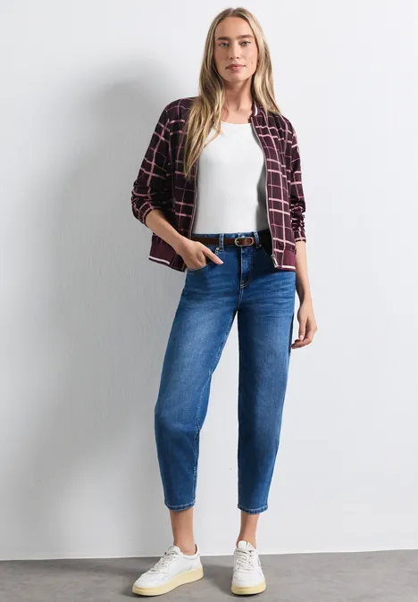 Frau mit kariertem Hemd, weißem Top, blauen Jeans und weißen Sneakers vor hellem Hintergrund