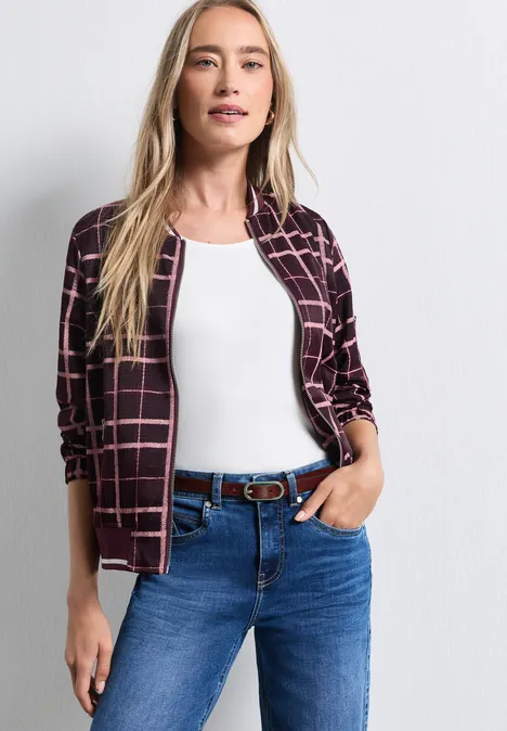 Junge Frau mit blonden Haaren trägt weiße Bluse, karierte Jacke und blaue Jeans mit Gürtel.