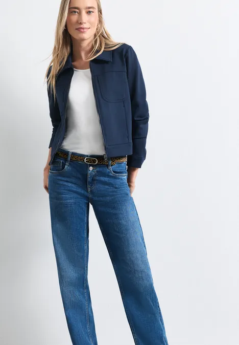 Vrouw in blauwe jas, wit shirt en spijkerbroek met riem voor een witte achtergrond.