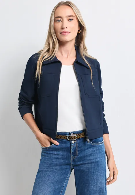 Blonde vrouw in donkerblauw jasje, wit topje en spijkerbroek met riem, handen in de zakken.