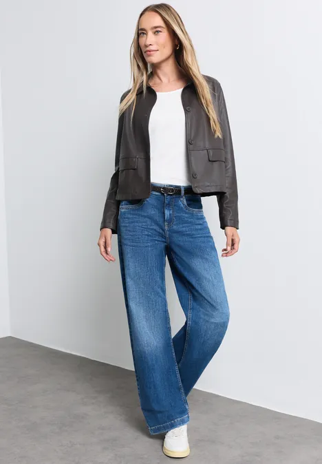 Frau mit blondem Haar, schwarzer Lederjacke, weißem Shirt, weiten Jeans und weißen Sneakern vor grauer Wand.