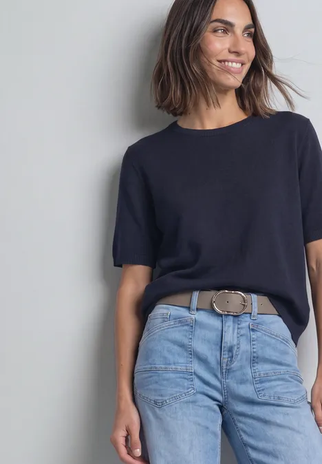 Gürtel mit dekorativer Schnalle Muddy Mocha Frau lehnt lächelnd an grauer Wand, trägt schwarzes T-Shirt, helle Jeans und auffälligen Gürtel.