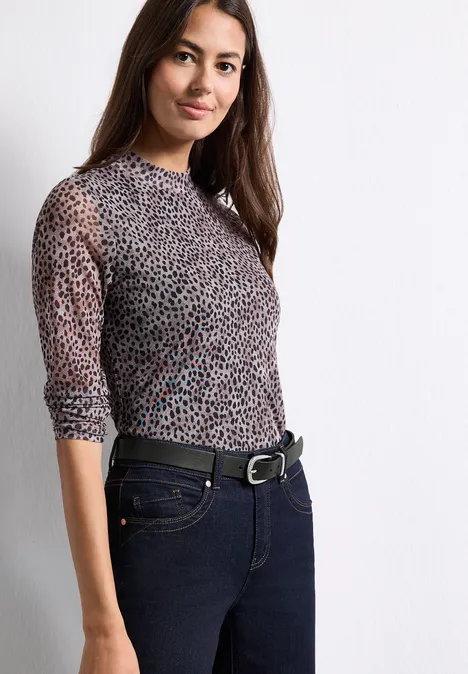 Frau in semi-transparentem Leopardenmuster-Shirt und dunkler Jeans vor weißem Hintergrund.