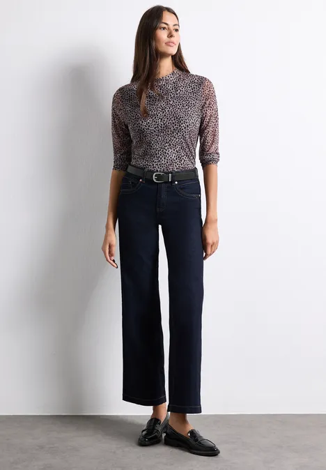 Frau in gemustertem Oberteil und weiten, dunklen Cropped-Hosen, schwarze Loafer.