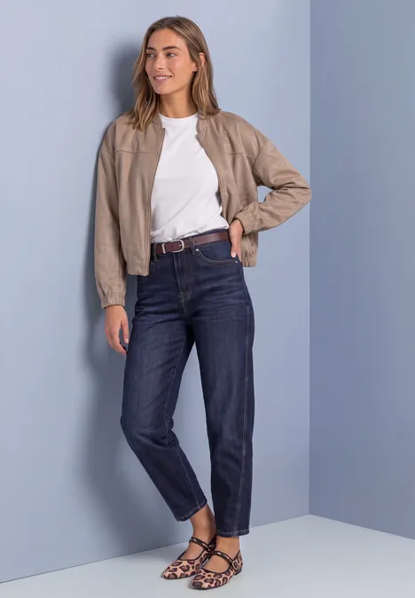 Une jeune femme porte une veste beige, un t-shirt blanc, un jean foncé et des sandales à motifs devant un mur bleu.
