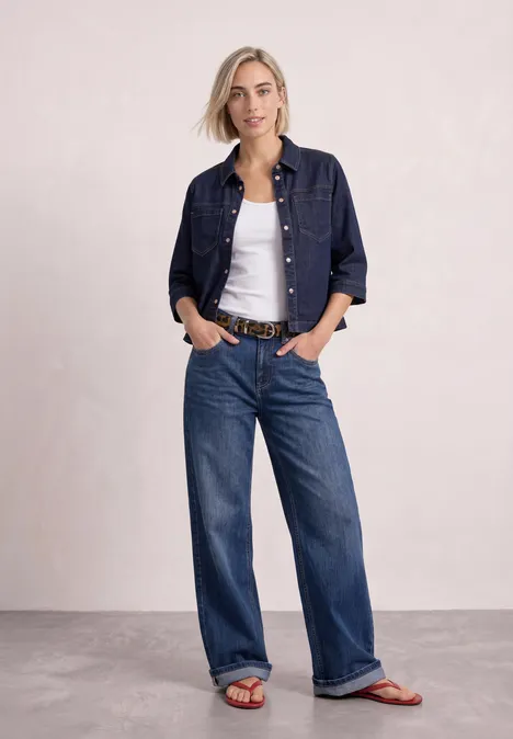 Riem met bont en luipaardprint cognac Frau in Jeansjacke, weißem Top und weitem Jeans, orangenen Sandalen vor neutralem Hintergrund.