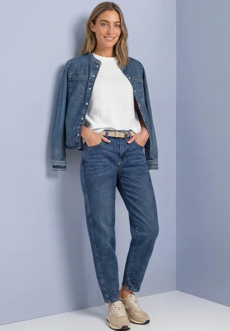 Velours Gürtel mit Nieten barley beige Frau trägt lässiges Jeans-Outfit mit weißem Shirt und Sneakern, steht vor hellvioletter Wand und lächelt.