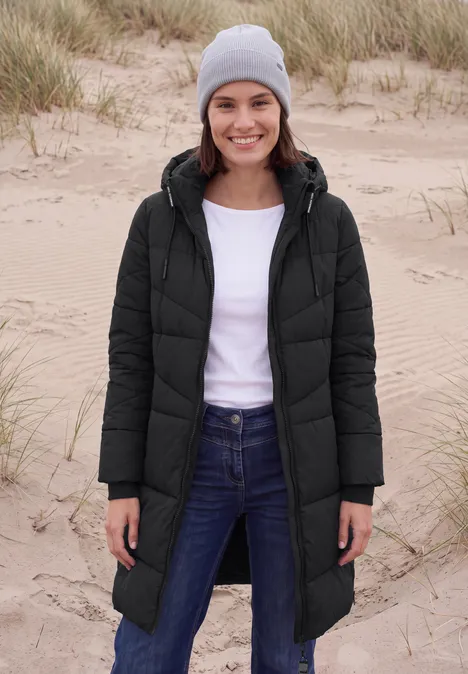 Steppmantel mit Kapuze und Reißverschluss Black Frau in schwarzer Steppjacke mit grauer Mütze steht am Strand vor Dünen.