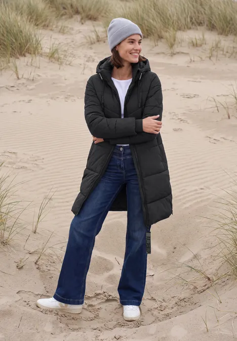 Steppmantel mit Kapuze und Reißverschluss Black Frau in langer, schwarzer Steppjacke mit Jeans, grauer Mütze und weißen Sneakern steht lächelnd am Dünenstrand.