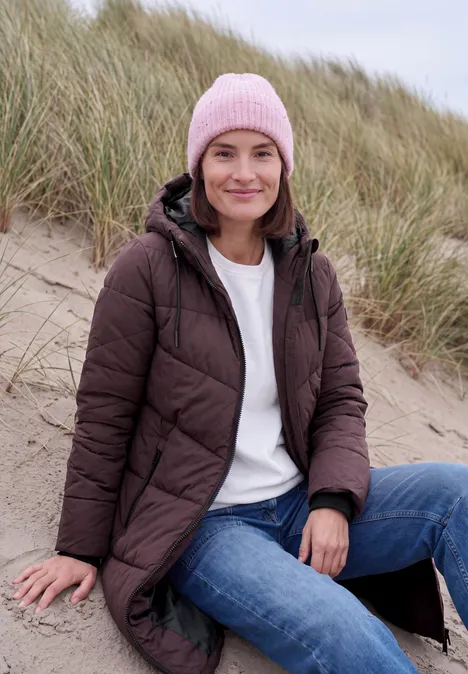 Steppmantel mit Kapuze und Reißverschluss berry red Eine lächelnde Frau sitzt am Strand zwischen Dünen, trägt eine braune Steppjacke, Jeans und eine rosa Mütze.