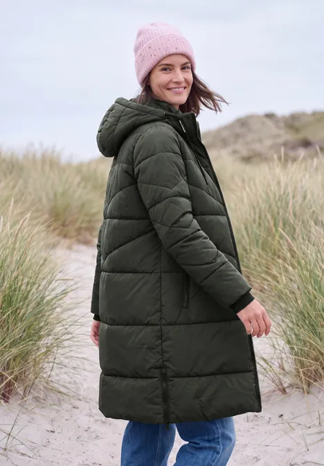 Femme souriante en manteau matelassé vert et bonnet rose sur un paysage de dunes.