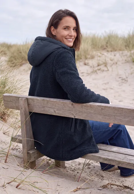 Eine lächelnde Frau sitzt auf einer Strandbank in den Dünen, trägt einen dunklen Mantel und schaut über die Schulter.