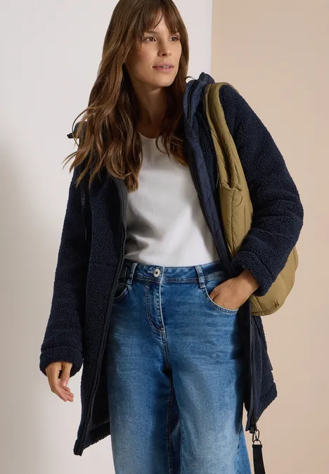 Eine Frau trägt eine dunkelblaue Jacke, ein weißes T-Shirt, blaue Jeans und eine olivgrüne Umhängetasche.
