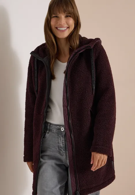 Strukturmix Teddy-Mantel mit Kapuze berry red Eine lächelnde Frau trägt einen dunklen, langen Teddy-Fleece-Mantel mit Kapuze und Reißverschluss.